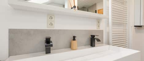 Une salle de bain moderne avec un meuble-lavabo blanc, deux robinets noirs, un distributeur de savon en bois et un miroir au-dessus du lavabo.