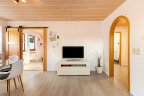 Un soggiorno moderno con televisione a schermo piatto su un mobile bianco, pavimento in legno e zona pranzo con tavolo e sedie in vetro.