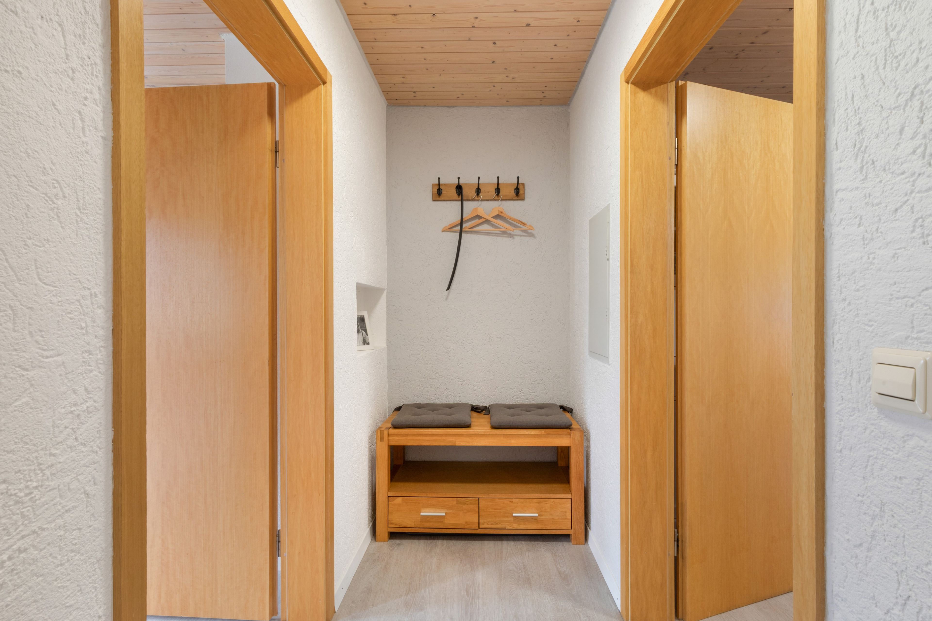 A narrow hallway with wooden doors on either side, a wooden bench with drawers, and a coat rack with hangers.