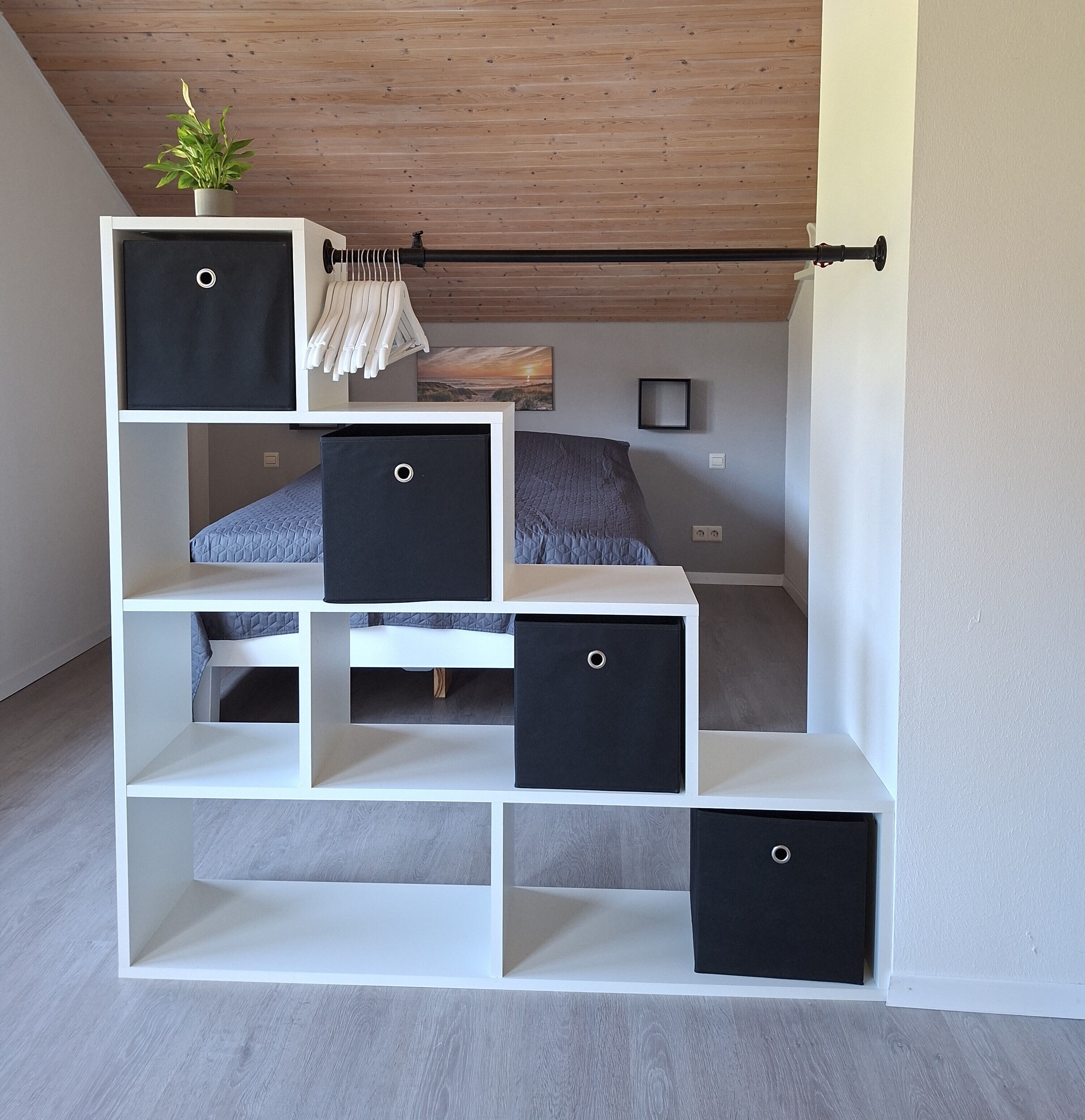 Una habitación pequeña con una cama litera, un estante blanco con cajas de almacenamiento negro y una pequeña planta en maceta.