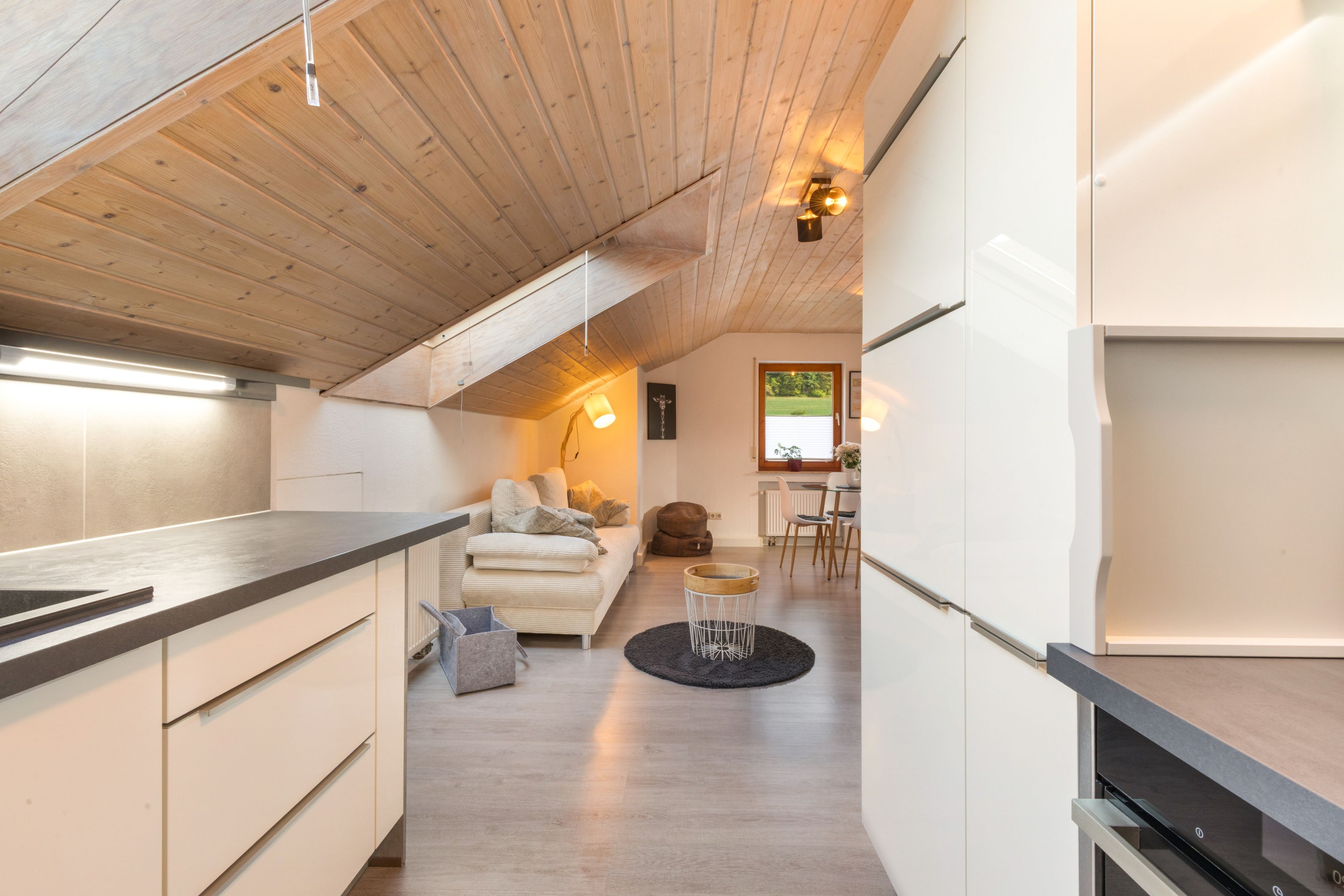 A modern kitchen with a wooden ceiling, a dining area with a table and chairs, and a living space with a sofa and a coffee table.