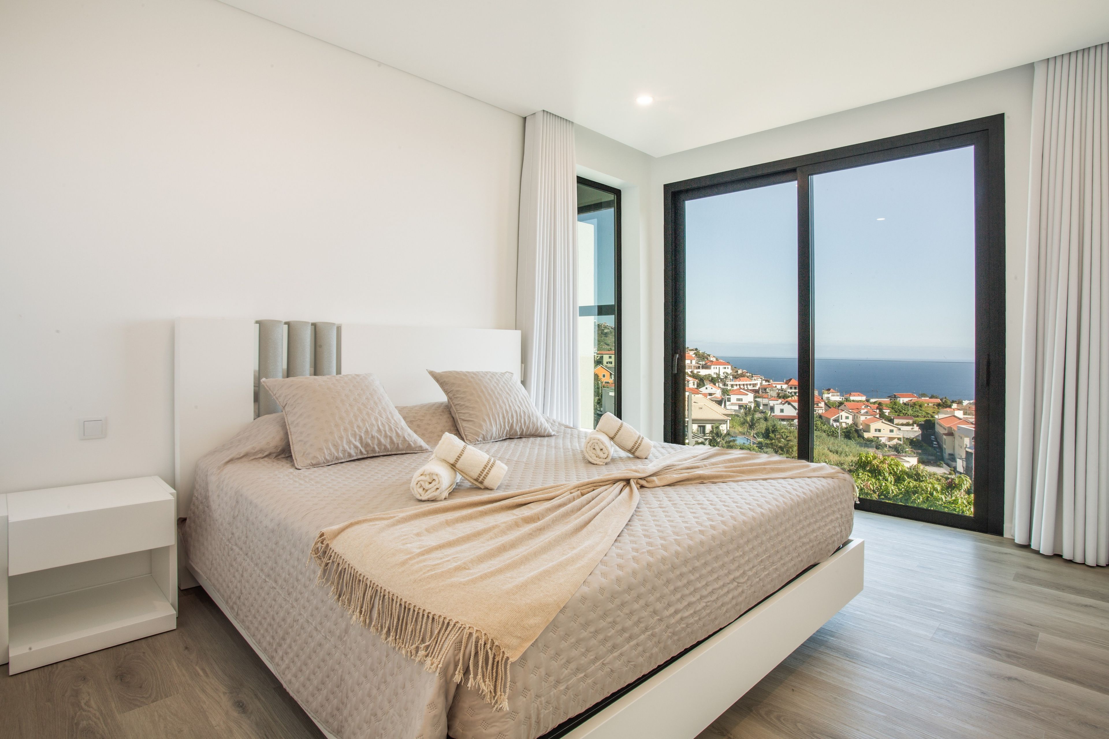 Une chambre moderne avec un grand lit, une table de chevet et une vue sur une ville côtière grâce à de grandes fenêtres du sol au plafond.