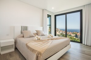 A modern bedroom with a large bed, a nightstand, and a view of a coastal town through floor-to-ceiling windows.