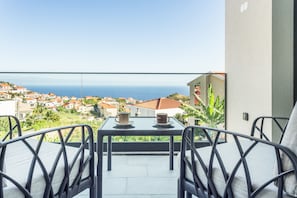 A balcony with a table and chairs, overlooking a coastal town with houses and the sea.