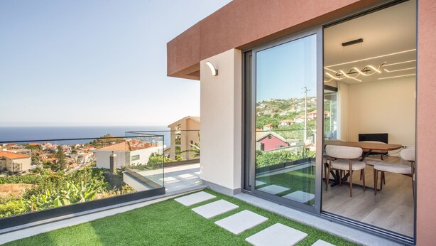 Un balcón moderno con puerta de cristal que da a un comedor, vistas despejadas al mar y un césped bien cuidado.