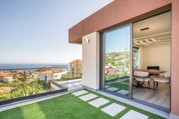 A modern balcony with a glass door leading to a dining area, a clear view of the sea, and a well-maintained lawn.