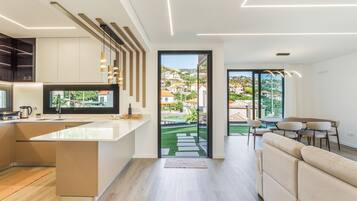 Une cuisine moderne dotée d’un grand îlot central, d’un coin repas avec un canapé et des chaises, et offrant une vue sur un quartier résidentiel grâce à une porte vitrée.
