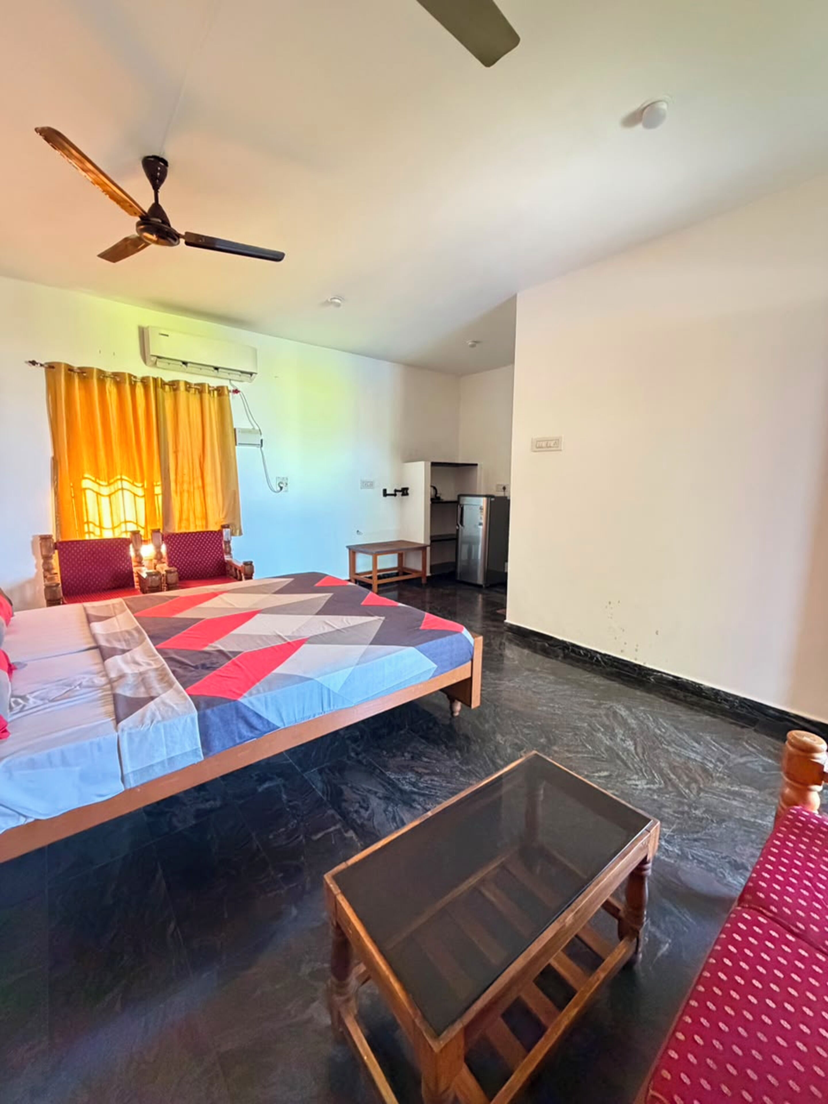 A bedroom with a bed, a small table, a ceiling fan, and a kitchenette in the background.