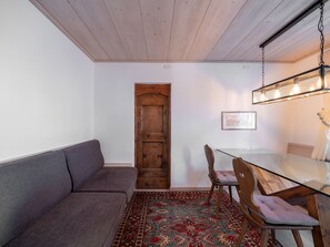 Een kamer met een houten plafond, een glazen tafel met stoelen, een grijze bank, een kamerbreed tapijt met patroon en een houten deur.