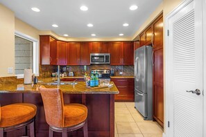 A kitchen with wooden cabinets, a stainless steel refrigerator, a microwave, and a granite countertop. - Kapalua Ridge | 2 Ocean View Villas, Sleeps 8 | Car Incl. w/ 6+ Nights | ML-4259 by KBM (Lahaina)