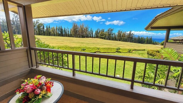 A balcony with a view of a golf course, a table with a floral arrangement, and a covered area with a roof. - Kapalua Ridge | 2 Ocean View Villas, Sleeps 8 | Car Incl. w/ 6+ Nights | ML-4259 by KBM (Lahaina)