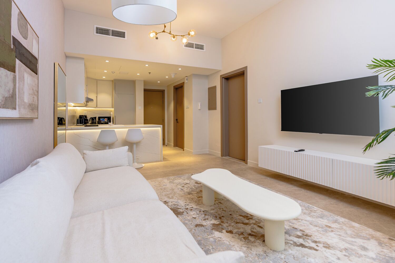 Una sala de estar moderna con un sofá, un banco, un televisor de pantalla plana y una cocina al fondo.