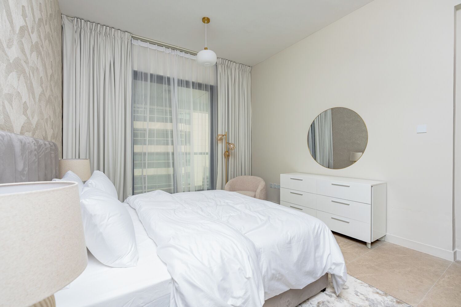 A bedroom with a bed, a bedside table, a lamp, a dresser, a round mirror, and a window with curtains.