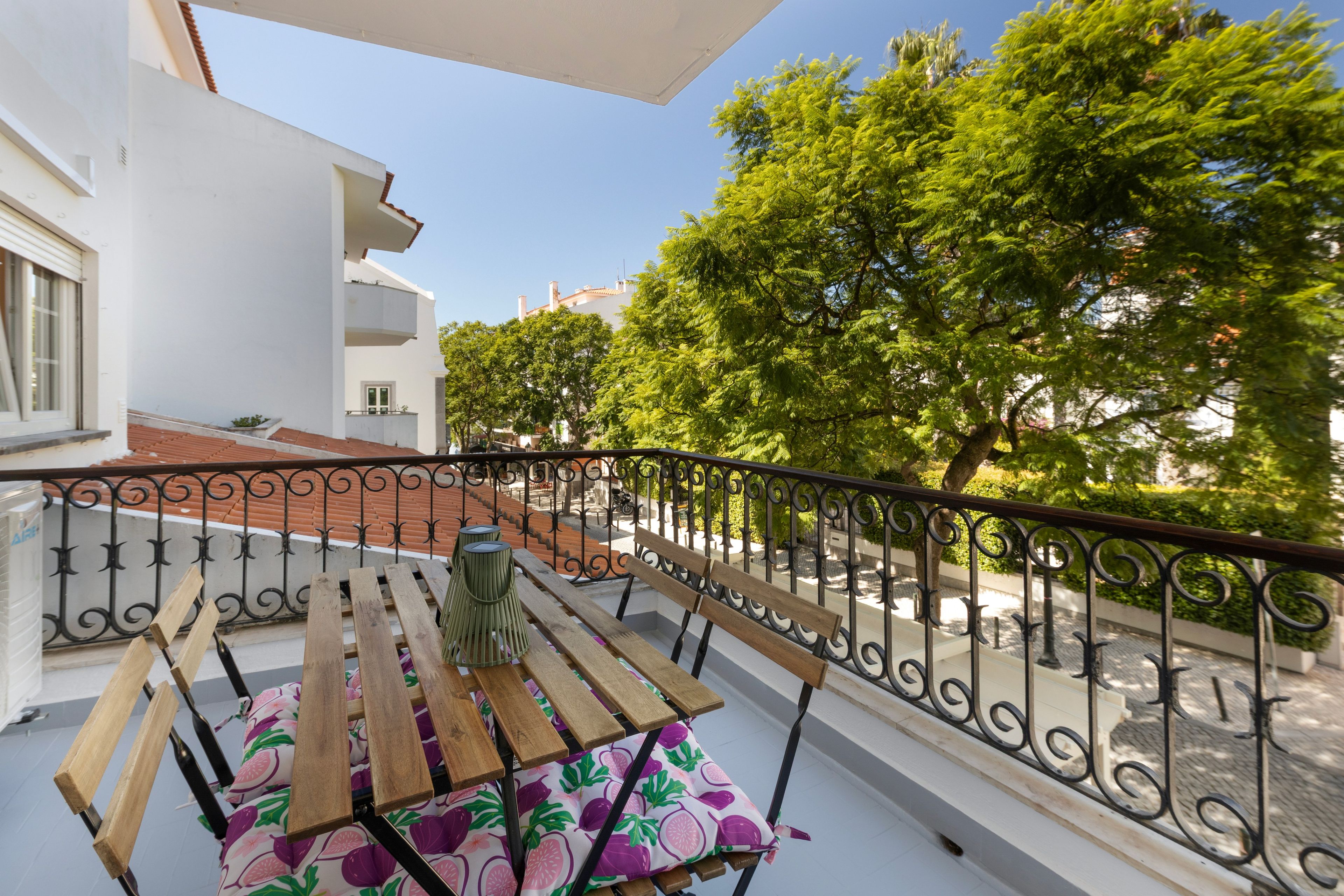A balcony with a wooden table and chairs, a metal railing, and a view of trees and buildings.