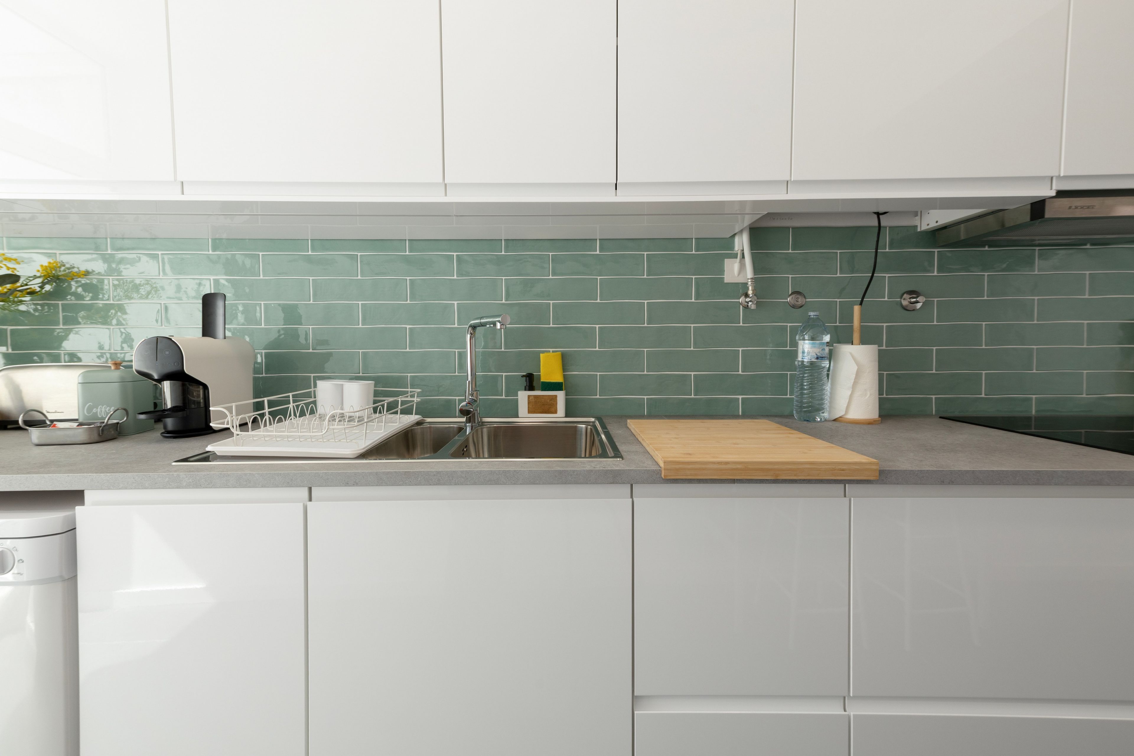 A modern kitchen with white cabinets, a stainless steel sink, and a green tiled backsplash.