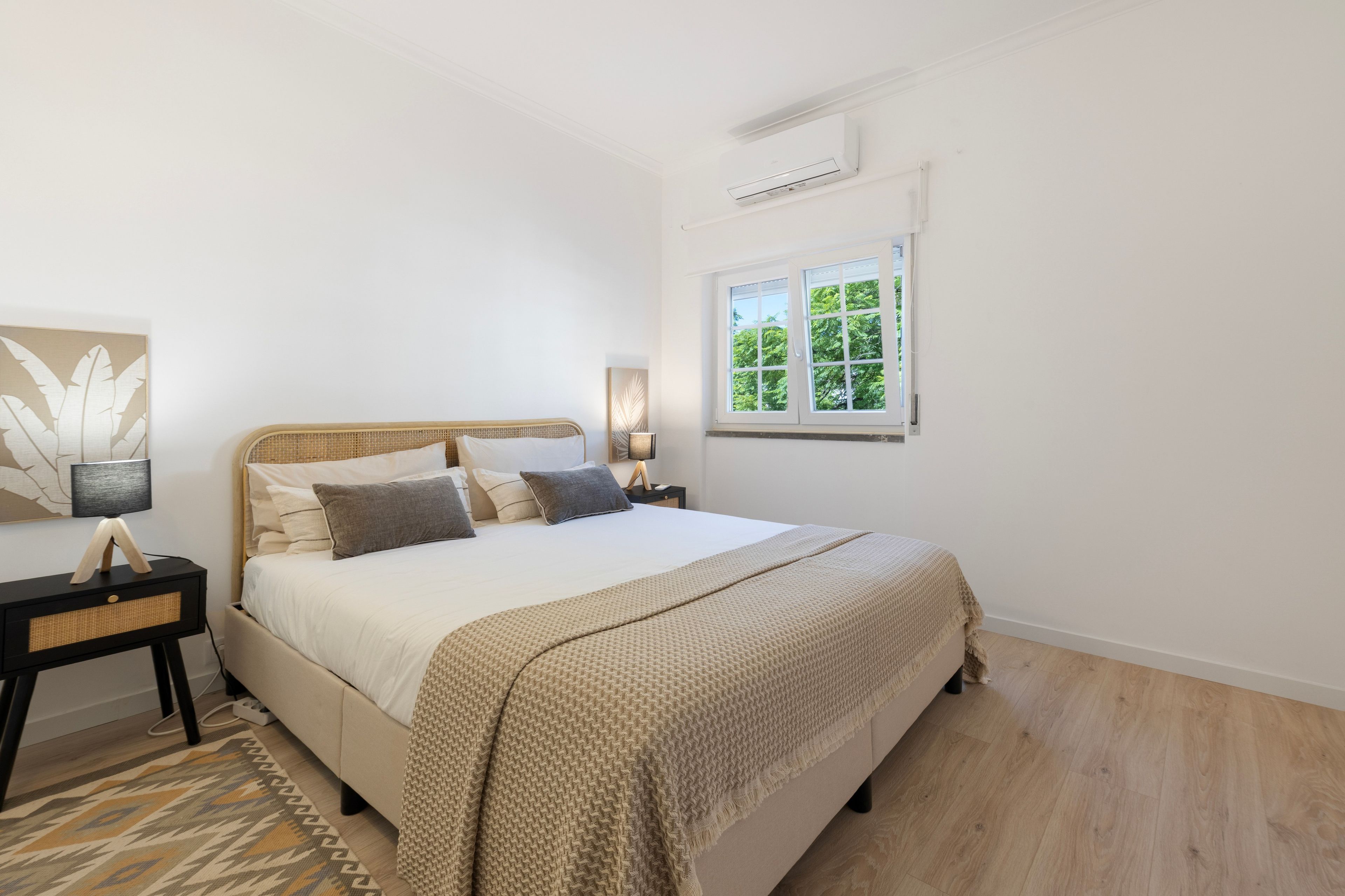 Un dormitorio con cama, mesitas de noche, una ventana con vistas a zonas verdes y un aire acondicionado de pared.