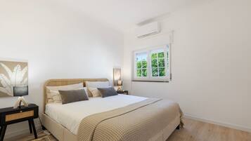 Une chambre à coucher comprenant un lit, des tables de chevet, une fenêtre donnant sur un espace verdoyant et un climatiseur fixé au mur.