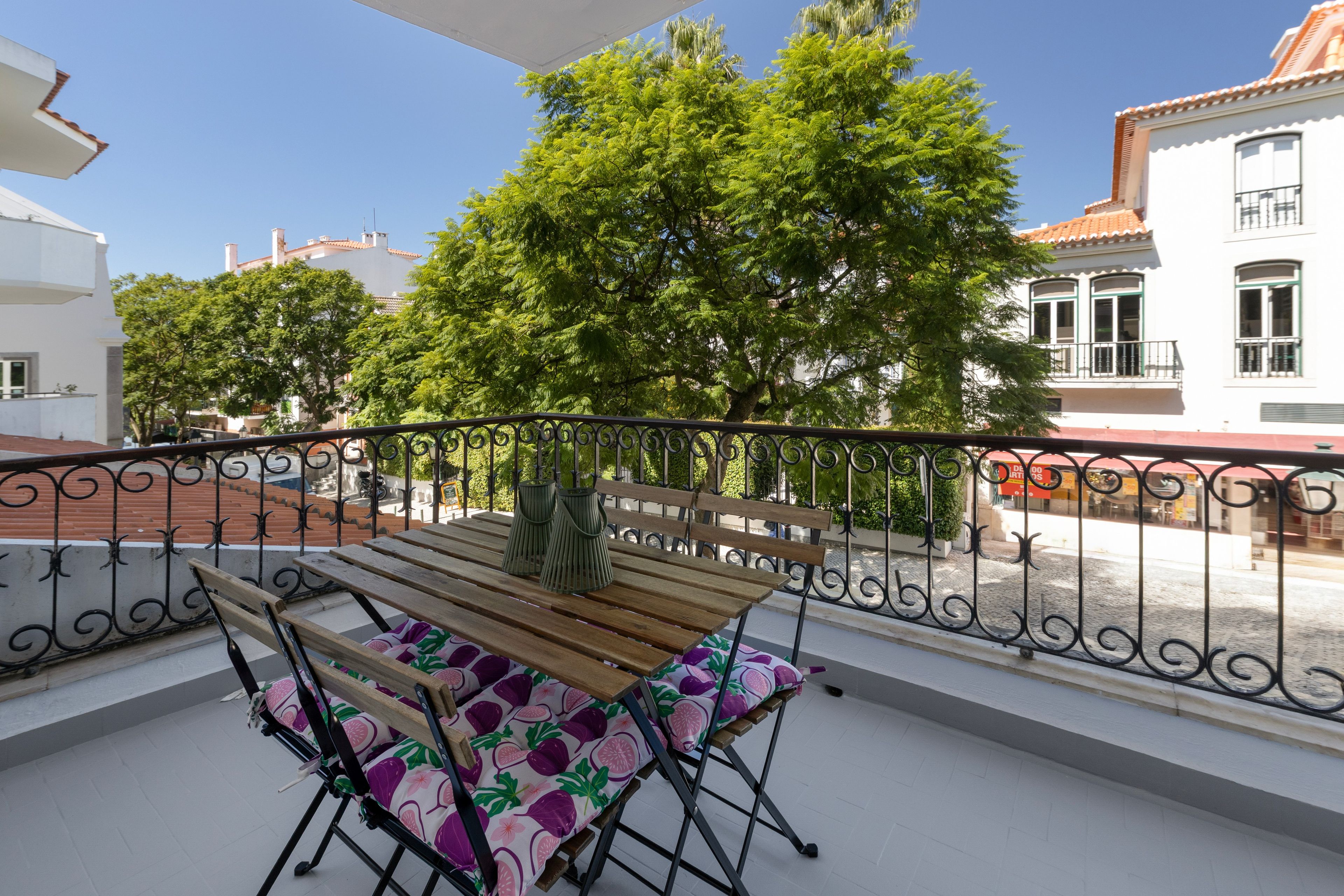 A balcony with a wooden table and chairs, a metal railing, and a view of trees and buildings.