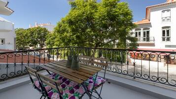 A balcony with a wooden table and chairs, a metal railing, and a view of trees and buildings.