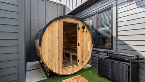 Une structure en forme de tonneau en bois, dotée d’une porte, d’un petit espace extérieur aménagé pour s’asseoir et d’une fenêtre avec des stores.