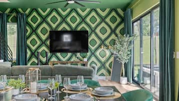 A dining area with a large table set for six, green upholstered chairs, a ceiling fan, and a flat-screen TV mounted on the wall.