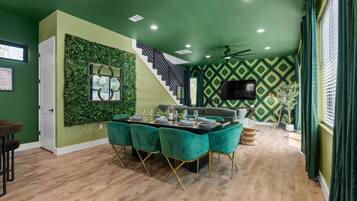 Una sala de estar moderna con una pared verde decorativa, una escalera, una zona de comedor con una mesa preparada para cuatro personas y un televisor de pantalla plana montado en la pared.