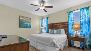 Quarto com cama, escrivaninha, ventilador de teto e janela com cortinas.