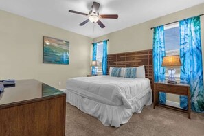 Ein Schlafzimmer mit Bett, Schreibtisch, Deckenventilator und einem Fenster mit Vorhängen.