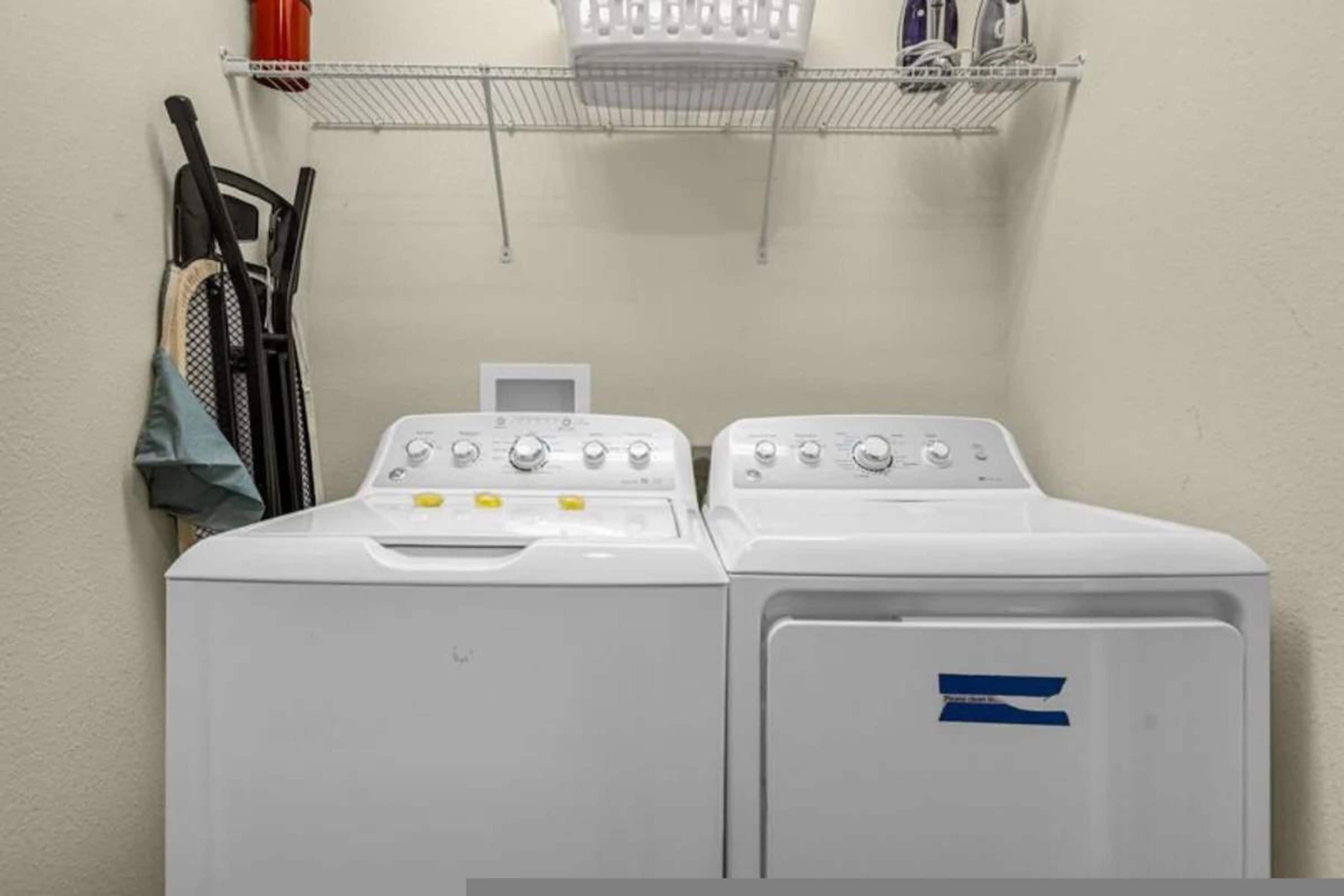 A laundry area with two white washing machines, a drying rack, and a shelf with various items.