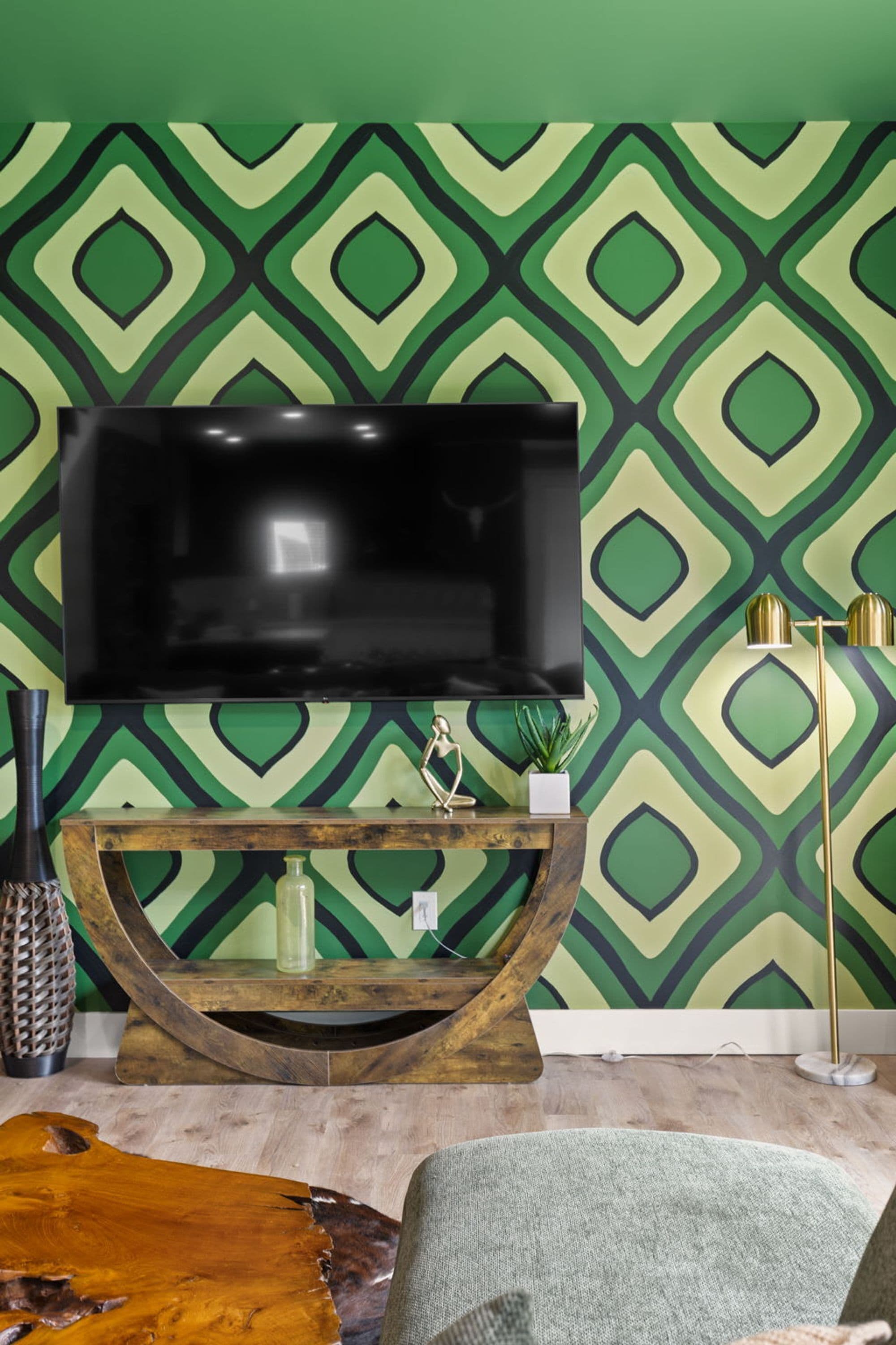 Un salon moderne avec un grand téléviseur à écran plat fixé sur une console en bois, un mur vert à motifs et un lampadaire.