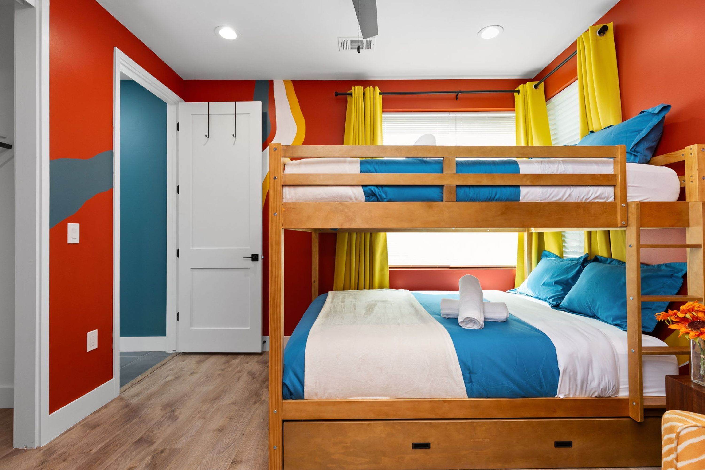 Uma cama de solteiro com estrutura de madeira, cabeceira branca e roupa de cama azul. Há uma toalha branca na cama, e cortinas amarelas são visíveis.