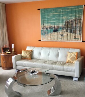 A living room with a white leather sofa, two patterned throw pillows, a glass coffee table, and a large abstract painting on the wall. - 2-bedroom condo in awesome Fort Lauderdale with AC and gym access (Fort Lauderdale)