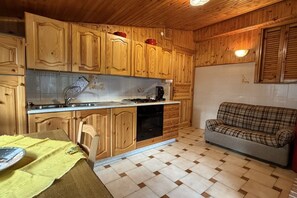 Eine Küche mit Holzschränken, einem Esstisch mit gelber Tischdecke und einem karierten Sofa.