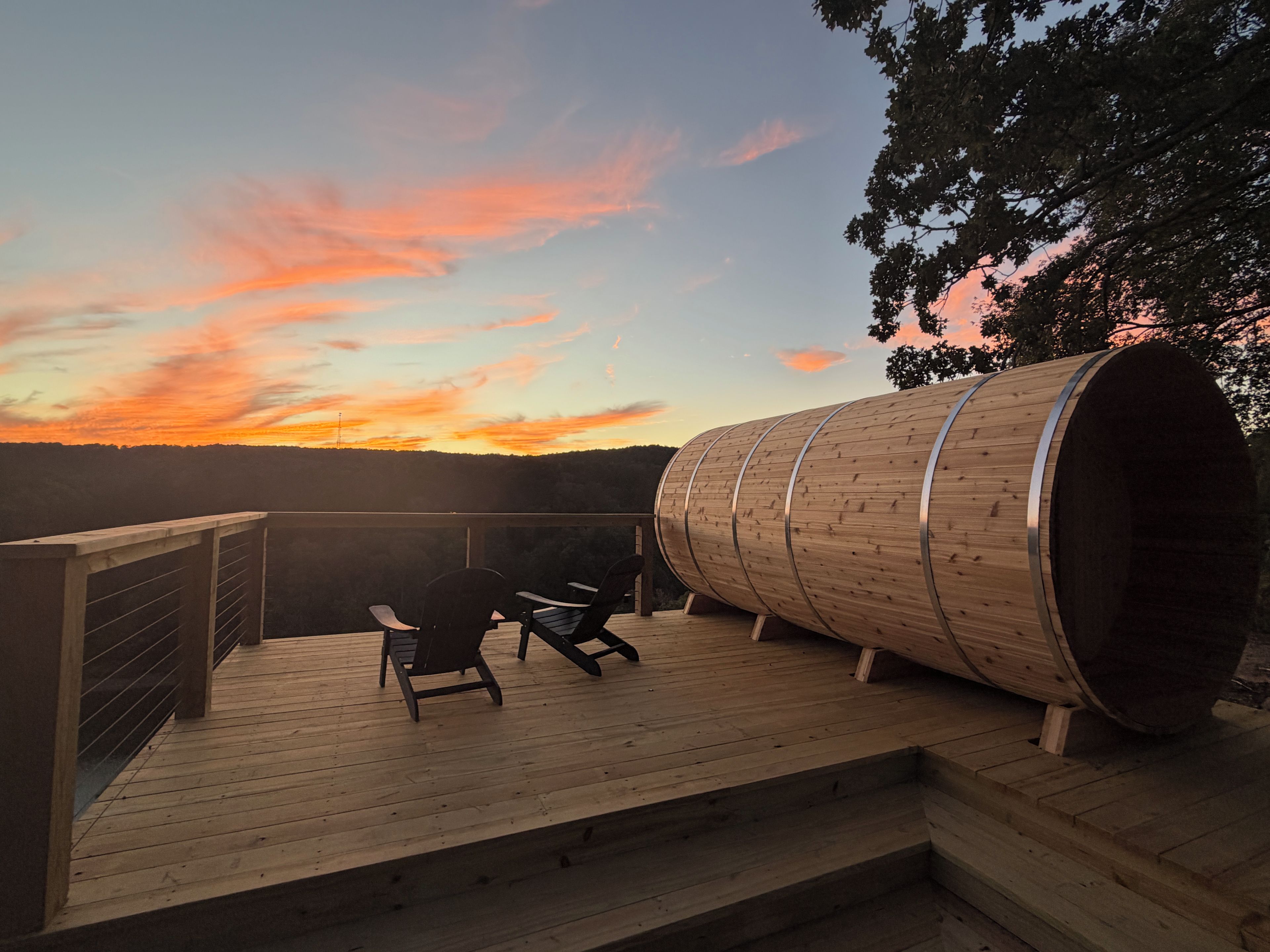 A wooden deck with two chairs and a large cylindrical wooden structure, likely a sauna, overlooking a sunset.