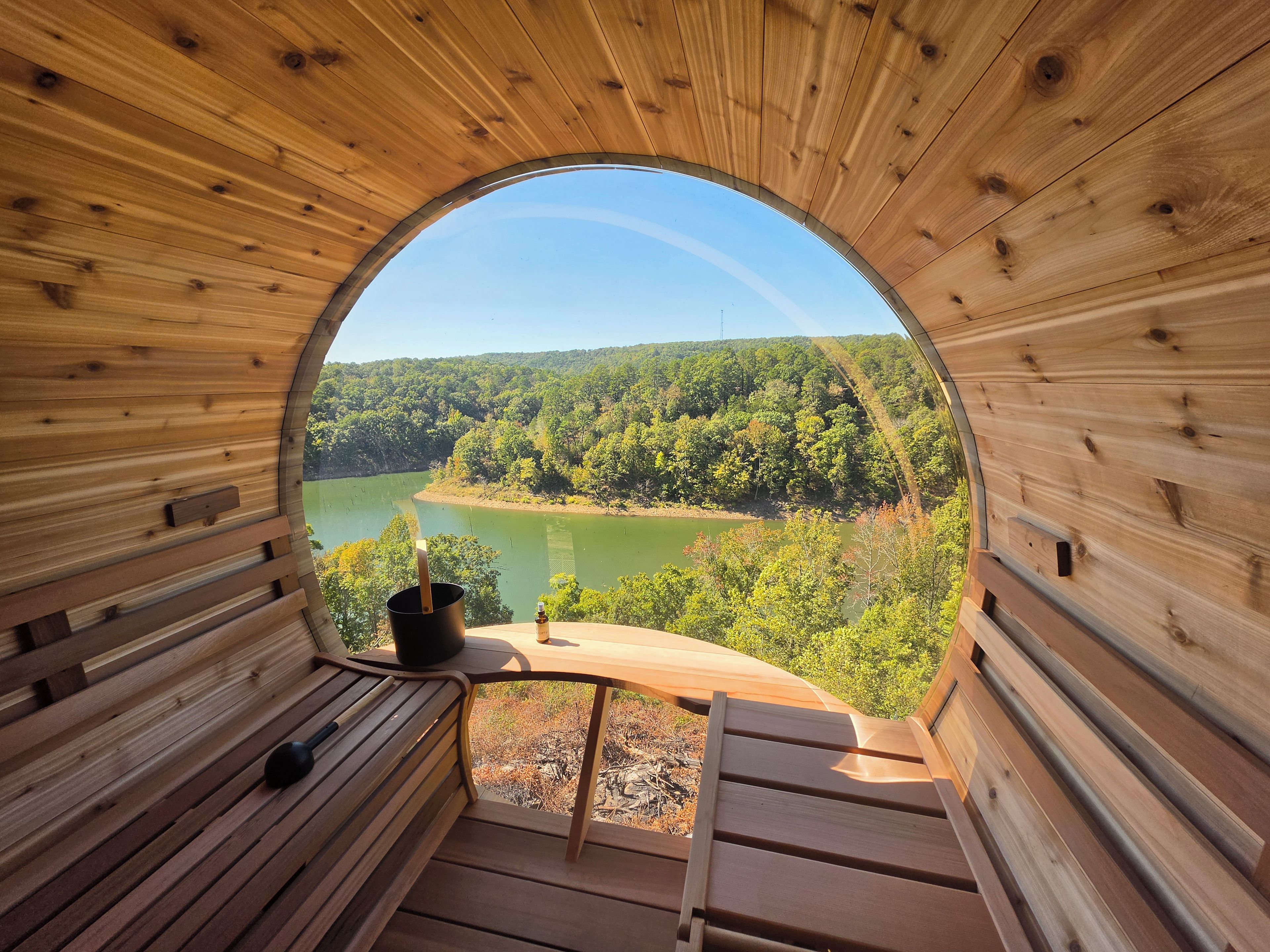 Una estructura circular de madera con un banco y una mesa, con vista a un río rodeado de bosque.