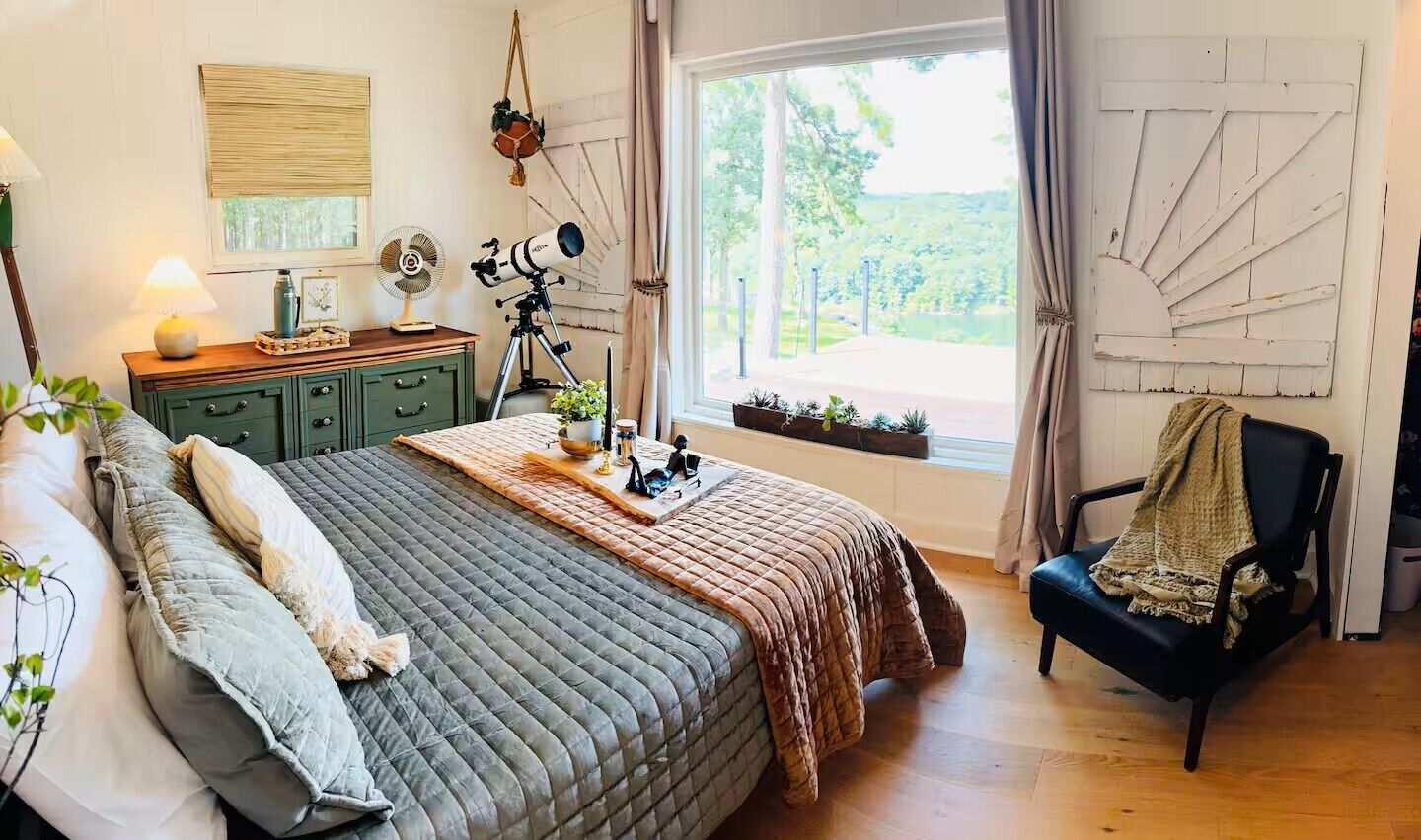 Ein Schlafzimmer mit einem Bett, einem Schreibtisch, einem Stuhl, einem Teleskop und einem Fenster mit Blick auf Grünflächen.
