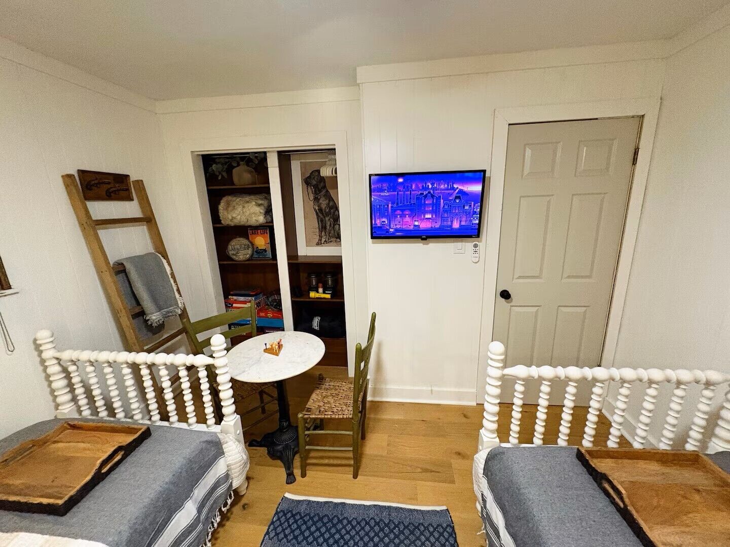 A cozy room with a white door, a television mounted on the wall, a small round table, two white chairs with wooden legs, a bookshelf, and a ladder.