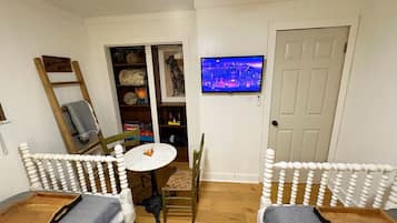 A cozy room with a white door, a television mounted on the wall, a small round table, two white chairs with wooden legs, a bookshelf, and a ladder.