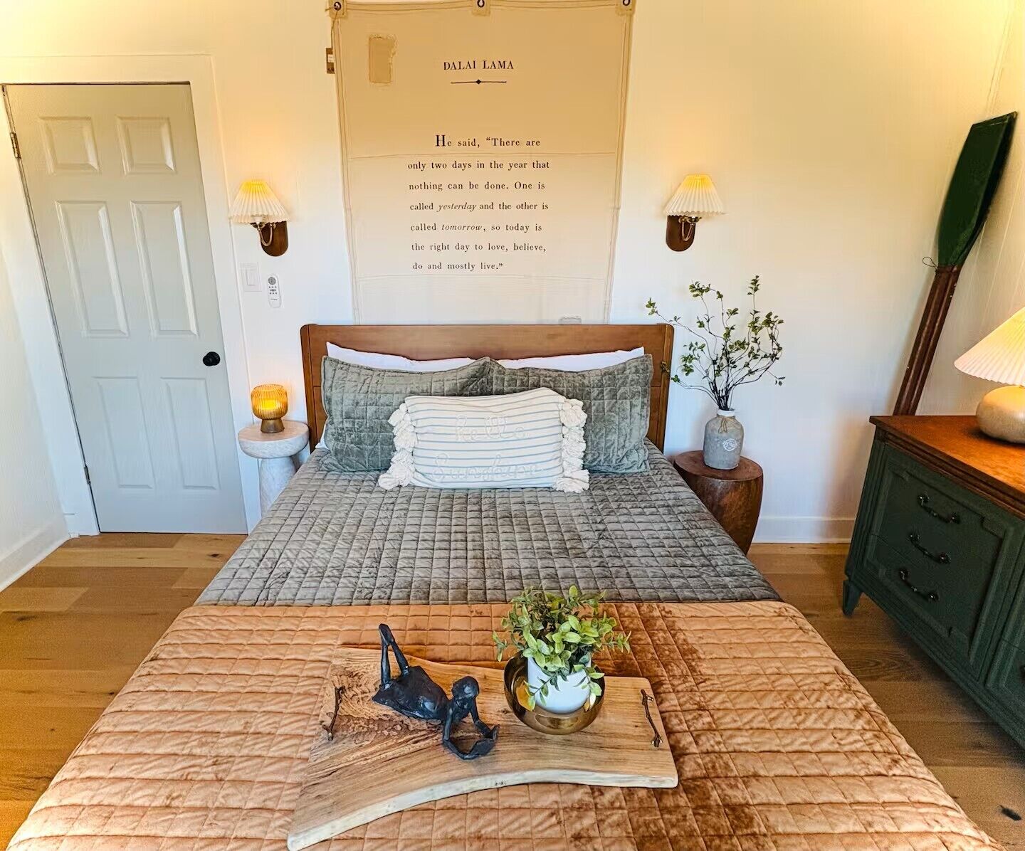 A bedroom with a bed, bedside lamps, a wall-mounted lamp, a bedside table with a vase, a dresser, and a poster on the wall.
