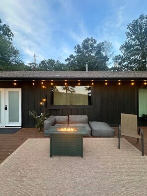 A covered outdoor seating area with a fire pit table, a cushioned bench, and a view of the surrounding trees.