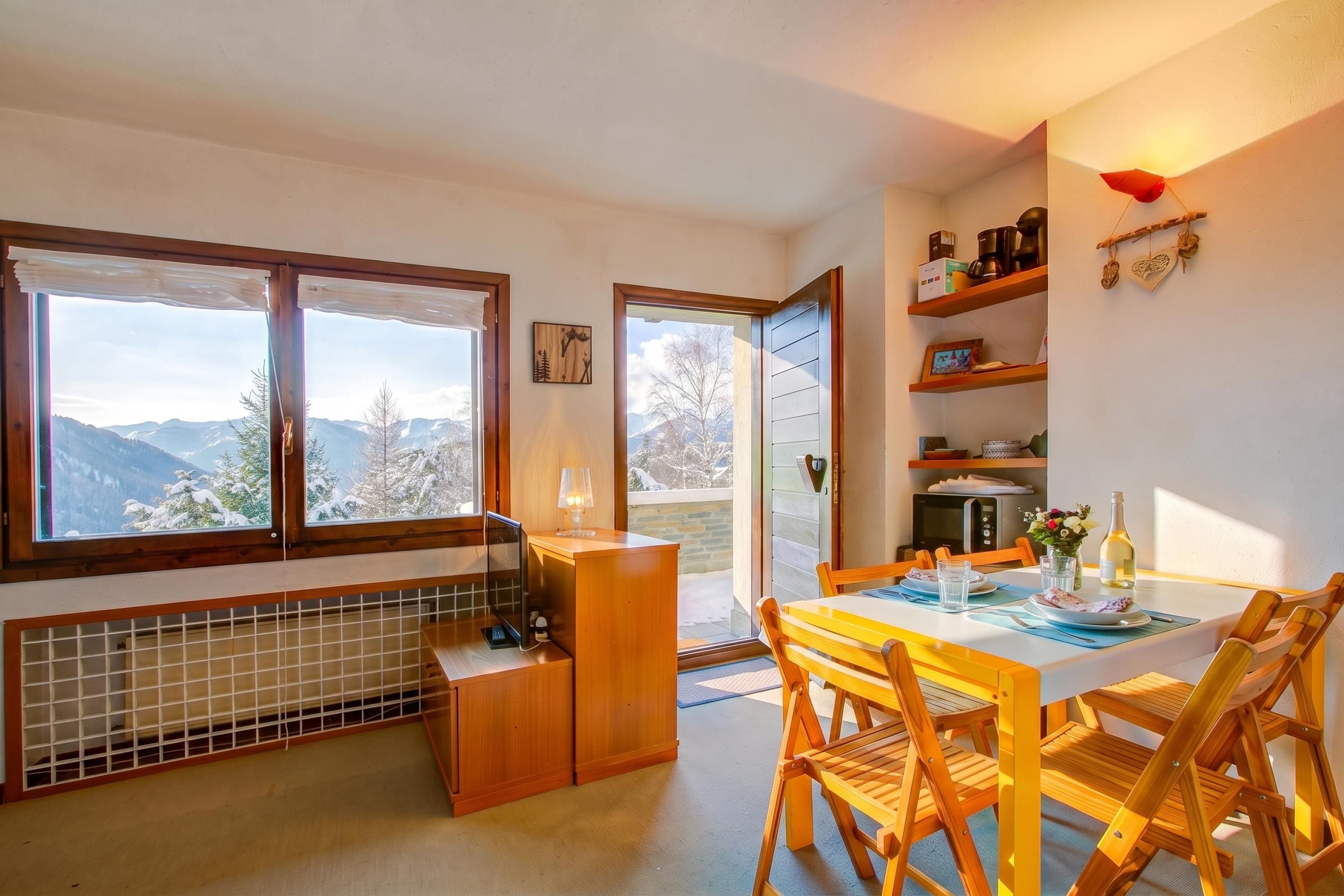 Een eetruimte met een tafel en stoelen, uitzicht op bergen door grote ramen, en een keuken met een fornuis en kastjes.