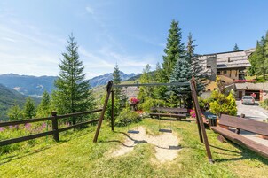 Un columpio de madera, un banco y un sendero pavimentado en un jardín con montañas al fondo.