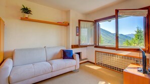 A room with a white sofa, a small table, a window with a view of mountains, and a radiator. - Appartamento I Larici (Sauze di Cesana)