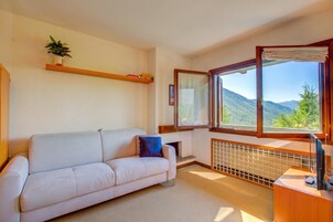 Una habitación con un sofá blanco, una mesita, una ventana con vistas a montañas y un radiador.