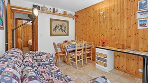 A cozy living room with a patterned sofa, a dining area with a table set for four, and a kitchen area with wooden paneling and a coffee machine. - Appartamento La Radura (Claviere)