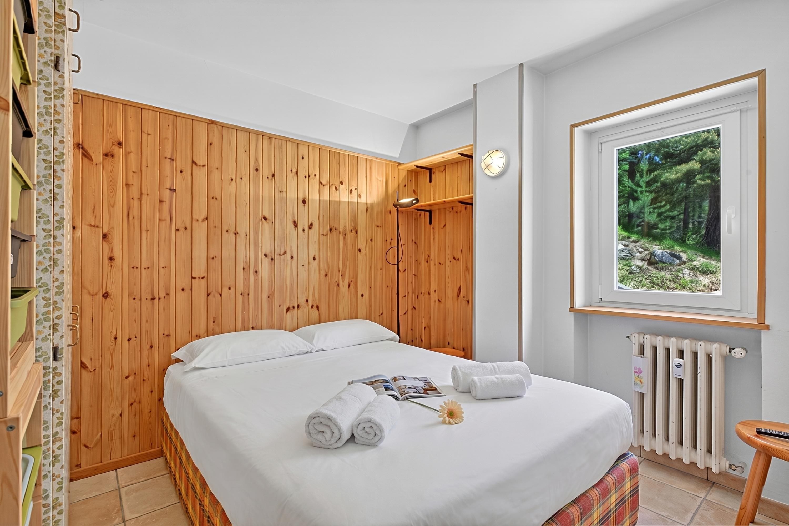 A bedroom with a wooden panel wall, a bed with white linens, a window with a forest view, and a radiator.