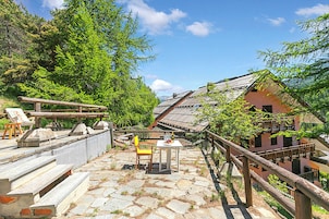 Una escalera de piedra conduce a una terraza de madera con una mesa y sillas, rodeada de abundante vegetación y edificios tradicionales.