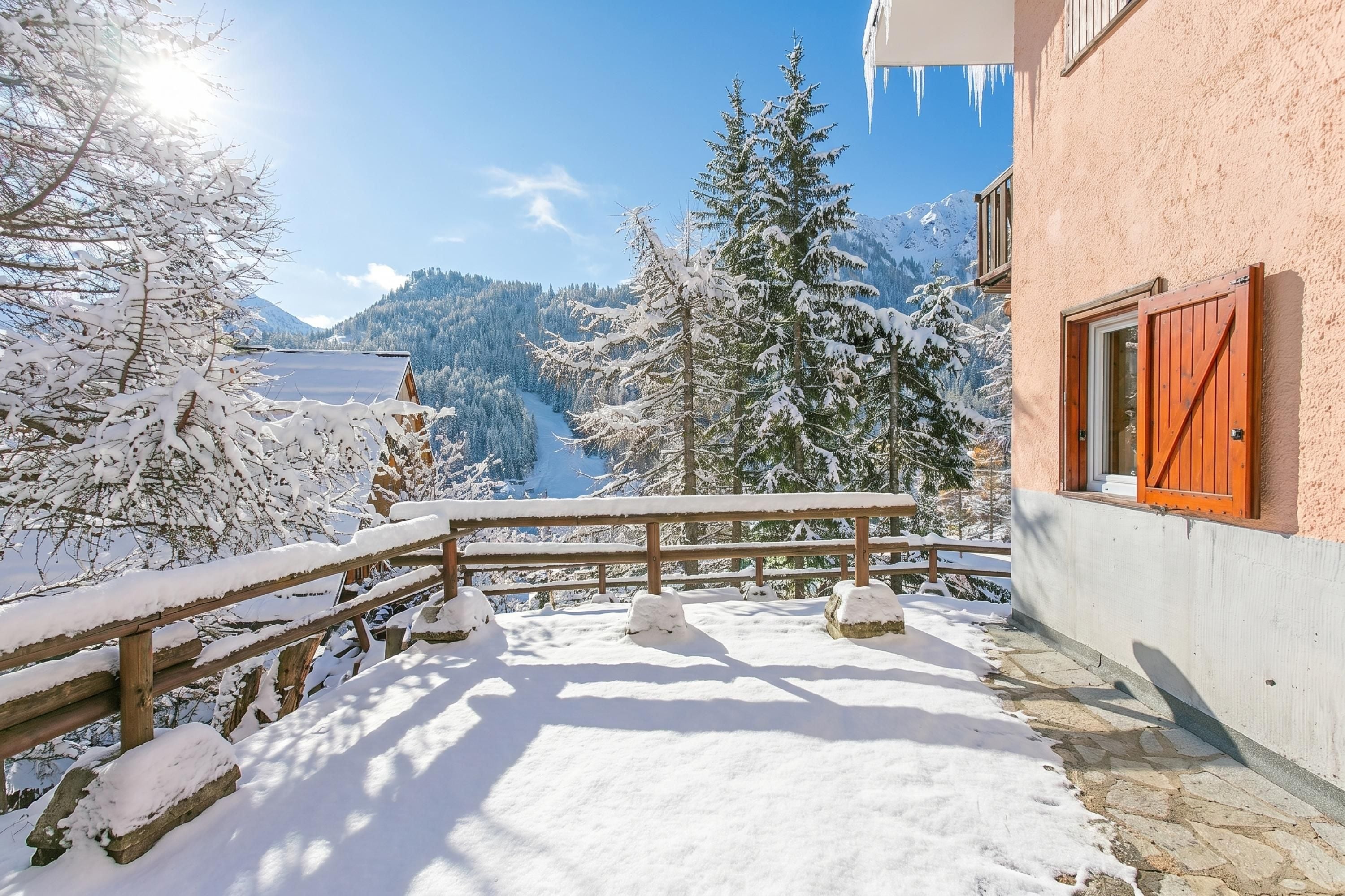 A snow-covered balcony with a wooden railing, a stone pathway, and a view of snow-laden trees and mountains.