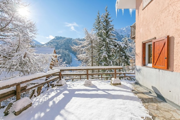 A snow-covered balcony with a wooden railing, a stone pathway, and a view of snow-laden trees and mountains. - Appartamento La Radura (Claviere)