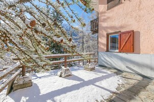 A snow-covered outdoor area with a wooden bench and a stone table, a building with a pink wall and a window, and a mountain in the background.